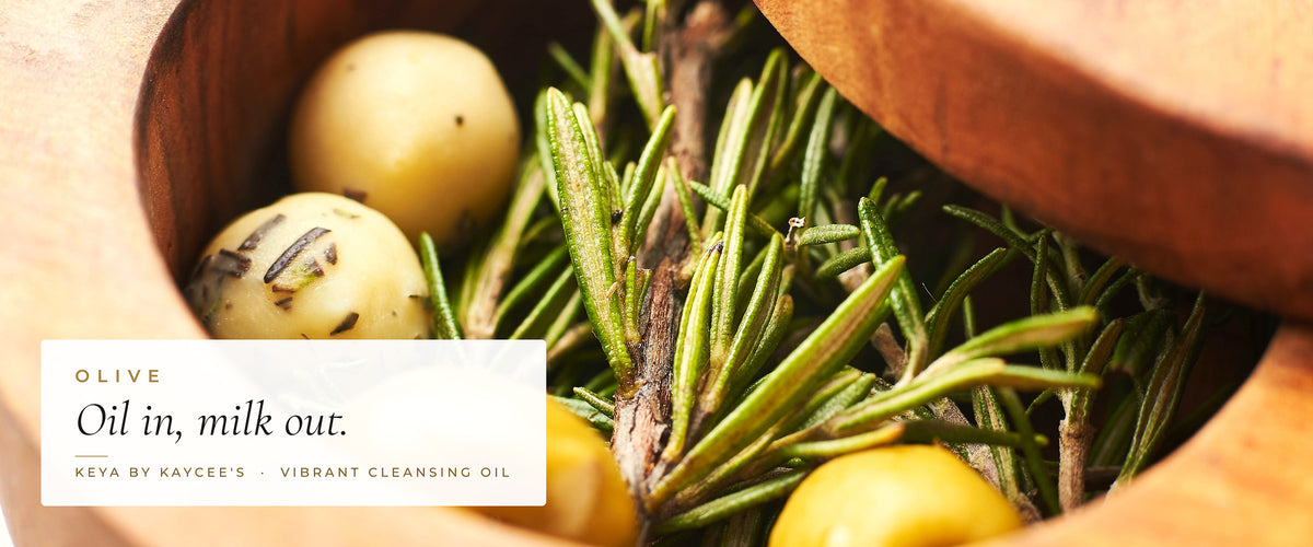 Olives and rosemary in a wooden bowl with the words Oil in, milk out, for KEYA Vibrant Cleansing Oil